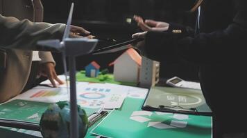 Two people standing near a table with papers and a wind turbine video