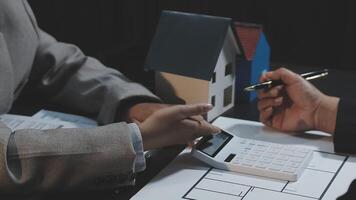 Two people are working on a house model with a calculator video