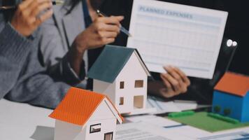 A group of people are sitting around a table with a house model video