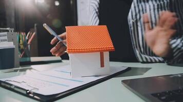 A real estate agent is holding a house model in front of a laptop video