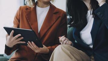 Two women in business attire sitting on a couch looking at a tablet video