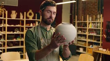 Happy smiling Caucasian salesman potter male man guy businessman artist small business owner holding pottery clay ceramic inspecting vase pot giving product to customer looking at camera in workshop video
