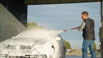 A man using a red pressure washer to apply foam on a car windshield video