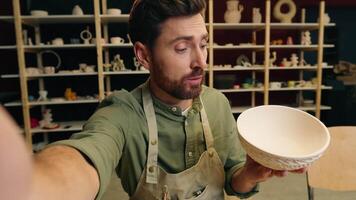 Talented Caucasian salesman potter male man guy businessman artist small business owner holding pottery ceramic bowl record blog on mobile phone talking looking webcam camera in workshop store video