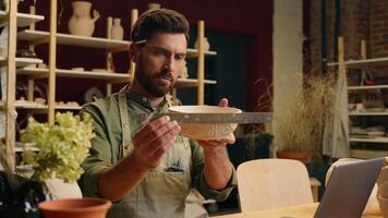 Serious Caucasian man businessman guy male small business owner seller potter measures clay bowl using ruler measure artwork calculate calculator laptop computer device indoors workshop pottery store video