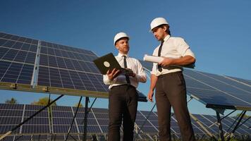 Two engineers in helmets and formal attire analyze data on a laptop and discuss the performance metrics of a solar power station video