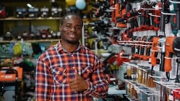 African-American man in a plaid shirt gives a thumbs-up while standing in a hardware store aisle filled with drills and power tools video