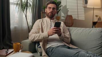 Man thoughtful dreaming Caucasian male homeowner tenant freelancer at home using mobile phone app online shopping in apartment smiling guy think on couch sofa relax browsing smartphone gadget addict video