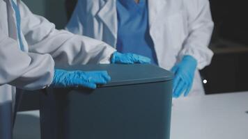 Two people in lab coats and gloves are holding a trash can video
