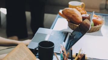 A woman is working on her laptop while sitting at a table with a basket of fruit and a video