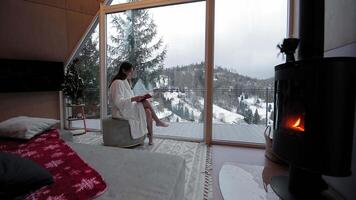 Woman in white robe reading by large window, enjoying scenic snowy mountain view and fireplace warmth. video