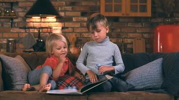 Two brothers of three and six years old are sitting on a sofa in the background of the kitchen, reading a book, looking at drawings and smiling video