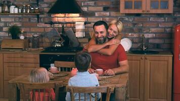 The wife hugs the husband sitting at the table with their two children. They gathered at dinner and are happy together and smiling. video