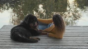 A young woman in a yellow jacket lies on the pier and strokes a large dog. Reflection of trees in the water. Camera movement forward video