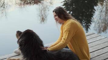 A young woman in a yellow jacket sits on a pier and strokes a large black dog. Reflection of trees in the water. Camera Move Right video
