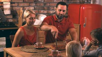 Happy friendly family dad mom and two children are sitting at a table in the kitchen on a cozy evening father cuts the cake that mom baked for dinner, the children look forward to trying a piece video