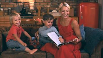 A beautiful young mother is reading a book to her two sons, three and six years old, sitting on a sofa against the background of the kitchen and smiling. video