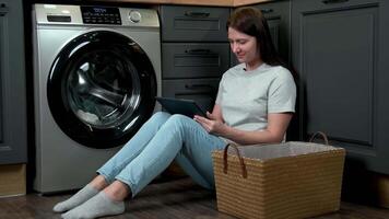 A woman uses a tablet while doing laundry while sitting next to a washing machine. video