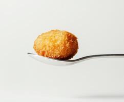 Crispy golden croquette on a silver spoon against a simple white background photo