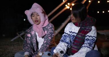 Handheld shot,Two Asian women in warm clothing sit roasting marshmallows over a campfire and chatting happily on a cold night. Winter weekend activity. video