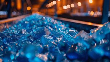 Sparkling blue glass fragments fill a conveyor belt in a factory illuminated by warm lights, showcasing the vibrant color and texture against the industrial backdrop at dusk. photo