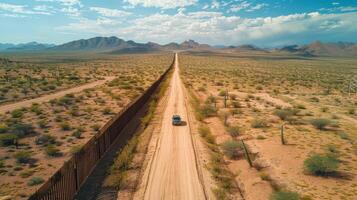A solitary vehicle travels along a long dirt road adjacent to a tall border wall in a vast desert landscape filled with cacti and scattered shrubs, surrounded by mountains in the distance. photo