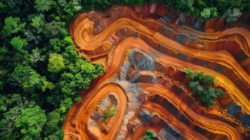 Heavy machinery reshapes the terrain in a tropical rainforest, leaving striking patterns of red soil and patches of green vegetation under a bright sky. photo
