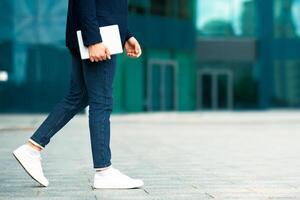 Unrecognizable young man in suit with modern digital tablet walking by street, cropped of IT manager going towards copy space, panorama, office buildings background. Technologies in business photo