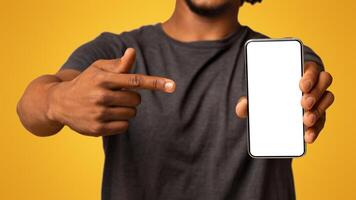 Smiling Black Male Pointing At Smartphone With Blank White Screen In His Hand, Cheerful African American Guy Recommending New Mobile App Or Website, Closeup Shot With Selective Focus, Mockup photo