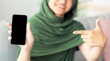 Unrecognizable Muslim teen girl in hijab pointing at cellphone with empty screen at home, mockup for design. Cheery Islamic adolescent offering smartphone with space for mobile app, closeup view photo