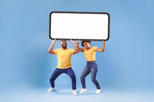 Millennial African American couple holding huge heavy smartphone with empty screen on blue studio background. Mockup for advertisement, mobile app or new website. Cellphone display template photo