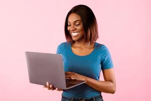 Happy black woman using laptop computer browsing internet posing standing over yellow studio background. People and gadgets, technologies, great website for freelancers concept photo