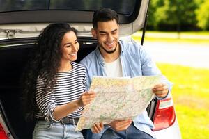 Happy millennial lovers tourists having break while car trip, cheerful young middle-eastern man and woman sitting on automobile trunk together, reading map, checking their location, empty space photo