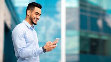 Positive middle eastern guy in formal outfit standing by modern business center, using smartphone and smiling, sending emails while having break, panorama with copy space, side view photo