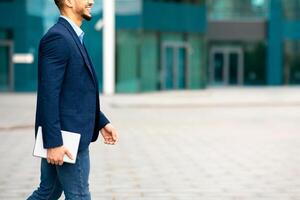 Unrecognizable young man in suit holding modern digital tablet walking by street, cropped of IT manager going towards copy space, panorama, office buildings background. Modern technologies in business photo