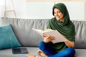 Happy teen girl in hijab sitting on sofa with textbook, reading, studying online from home, preparing for exam. Smart Eastern adolescent in headscarf learning remotely during coronavirus lockdown photo