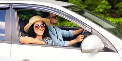 Smiling young lady in summer hat and sunglasses looking at camera while having car trip with her man, middle-eastern couple going vacation together, enjoying nice weather, side view photo