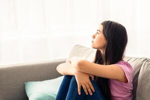 Teen depression concept. Stressed Indian teenage girl sitting on couch at home, looking out window, suffering from lack of communication, feeling upset and lonely during covid lockdown photo