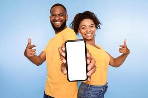 Use this new cool app. Positive African American couple pointing at smartphone with blank white screen, recommending mobile app, promoting your website, mockup. Selective focus photo