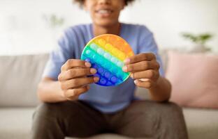 Cropped view of Afro teen guy playing with antistress POP IT toy indoors, closeup of hands. Unrecognizable black youth using sensory plaything at home. Stress relief, relax concept photo