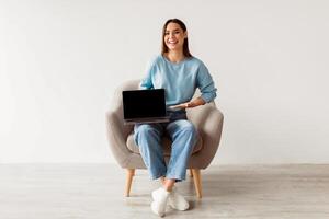 Check out this new website. Happy young lady sitting in armchair with laptop computer, showing blank screen with mockup, advertising your site design, full length portrait photo