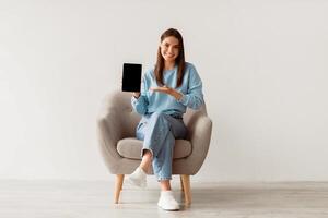 People and gadgets. Happy Caucasian lady showing laptop with blank screen for your website, presenting touch pad with mockup, sitting in armchair against white studio wall, full length photo