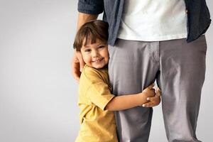 Father and son connection. Happy little boy embracing leg of his unrecognizable daddy, smiling to camera, feeling loved and safe, posing over grey wall background with empty space, panorama photo