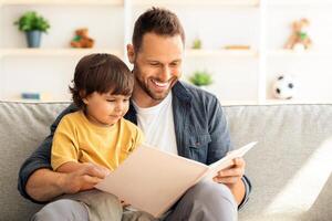 Weekend activities with toddlers. Cheerful carying daddy reading book to little son, baby boy enjoying fairy tale on dad's knees, sitting on sofa at home photo
