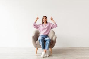 Excited Caucasian woman having idea and pointing two fingers up at copy space, sitting in armchair against white studio wall, mockup for your advertisement. Amazing offer photo