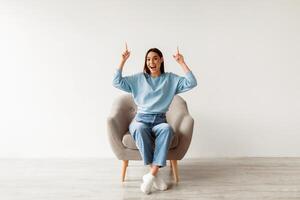 Amazing offer. Excited Caucasian woman sitting in armchair, pointing two index fingers up at empty space above her head against white studio wall, mockup for advertisement photo