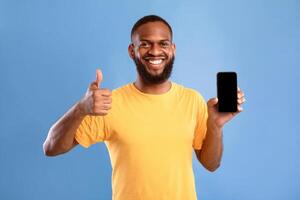 Advertisement for mobile application. Smiling young African American guy showing modern smartphone with blank screen, gesturing thumb up, recommending new app or website, mockup for design photo