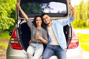 Emotional middle-eastern couple drinking coffee at country side, sitting on open auto trunk, embracing and gesturing, having coffee break while having car trip, enjoying vacation, copy space photo