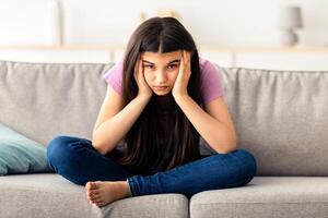 Irritated Indian teenage girl sitting on couch, feeling bored, suffering from lack of communication, being trapped at home during coronavirus lockdown, having teen depression, full length photo