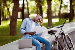 Upset black man with laptop computer and bicycle studying documents at park photo